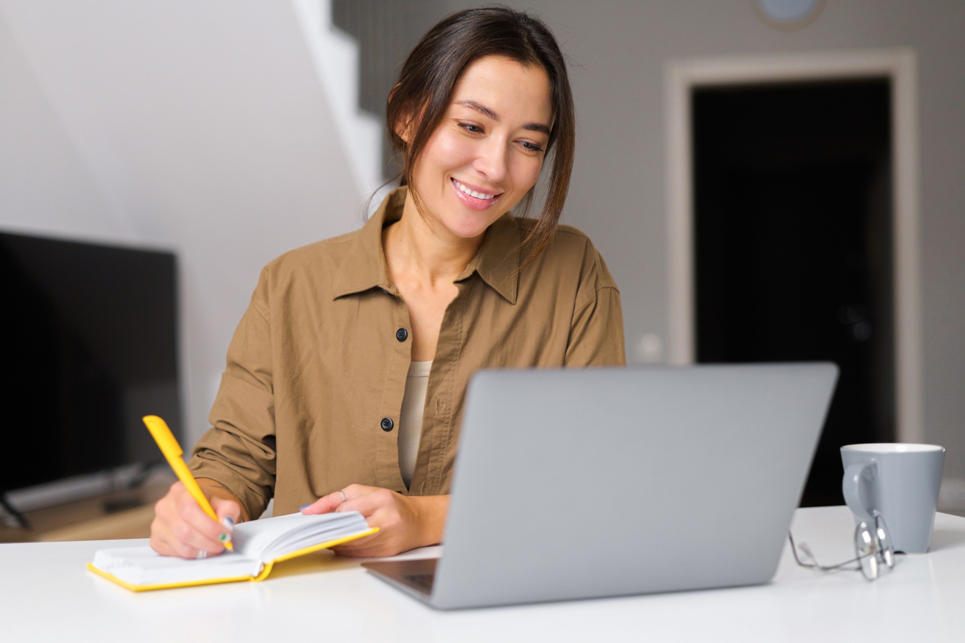 Smiling kind young woman works from home. She sitting at the kitchen table In front of the laptop and notebook